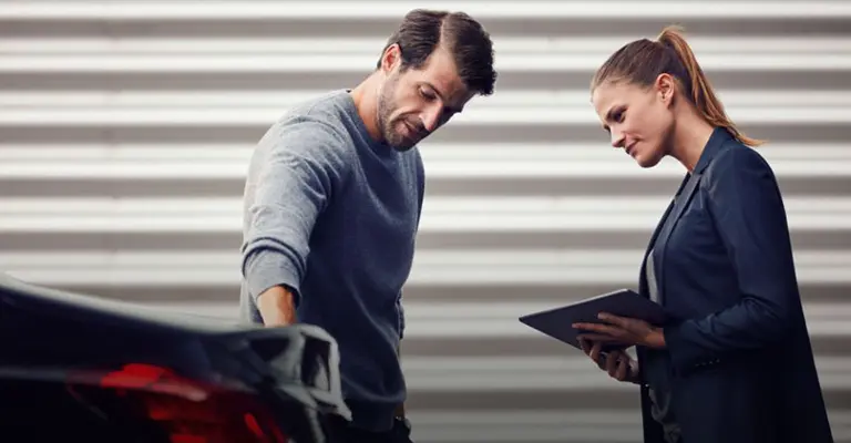 Ein Mann zeigt einer Frau mit Tablet Macken am Auto, da er den Verkehrsrechtschutz der Mercedes Creditcard in Anspruch nehmen will